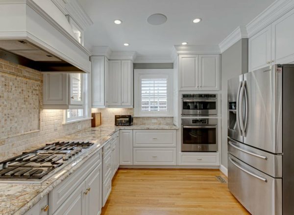 White kitchen with stainless steel appliances. Large refrigerator at right, ovens on left. Wooden floor with plantation shutters.